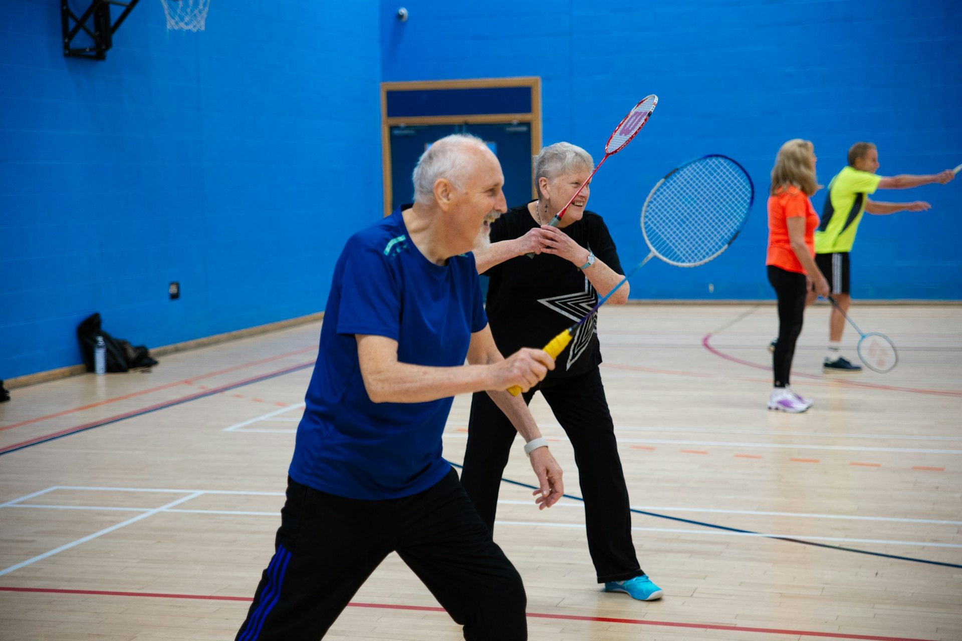 Ældre mand og ældre kvinde spiller grinende badminton i en idrætshal
