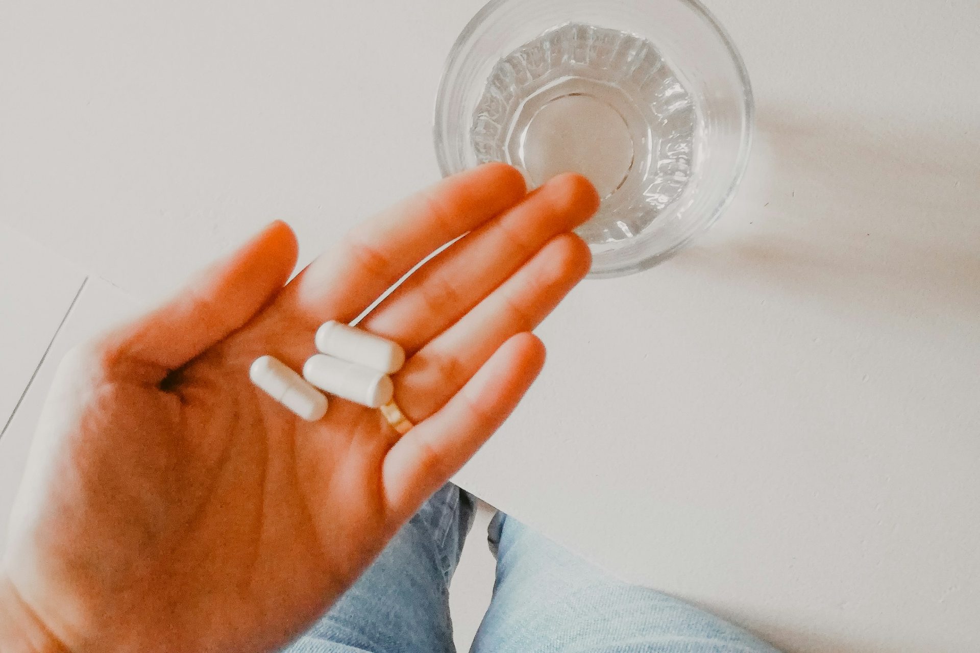 Hand holding three white capsules next to a glass of water