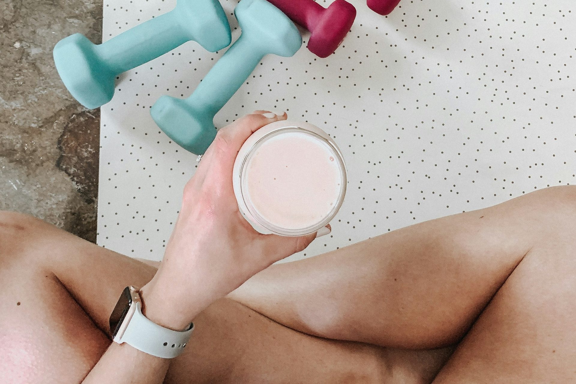 Woman sitting cross-legged holding a pink protein shake next to two pairs of dumbbells